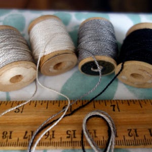 May include: Four spools of thread in silver, white, gray, and black sit on a wooden ruler. The spools are made of wood and have thread wound around them. The ruler is marked with inch measurements.