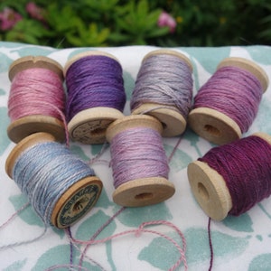 May include: Eight spools of thread in various shades of purple, pink, and blue. The spools are made of wood and have a vintage look.