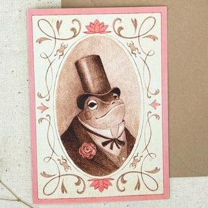May include: A greeting card featuring a watercolor illustration of a frog wearing a top hat and a bow tie. The frog is surrounded by a decorative frame with floral accents.