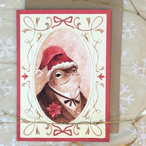 May include: A Christmas card featuring a watercolor illustration of a frog wearing a Santa hat and a bow tie. The frog is surrounded by a decorative frame with a poinsettia flower.