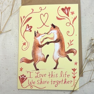 Card that says I love this life we share together in whimsical hand written letters with two foxes dancing. Card has a yellow background with red stylized flowers that boarder the orange dancing foxes. Art is in a semi-realistic children's book style