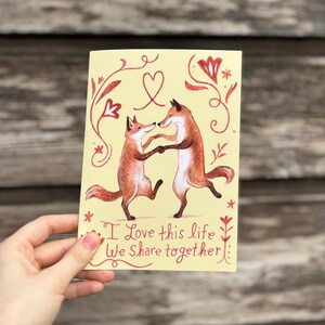 K&ouml;nnte beinhalten: Eine gelbe Karte mit zwei tanzenden F&uuml;chsen und einem roten Herzen dar&uuml;ber. Die Karte hat rote Blumenmuster und den Text "Ich liebe dieses Leben, das wir gemeinsam teilen".