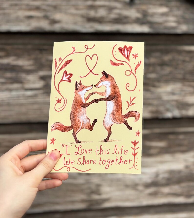 K&ouml;nnte beinhalten: Eine gelbe Karte mit zwei tanzenden F&uuml;chsen und einem roten Herzen dar&uuml;ber. Die Karte hat rote Blumenmuster und den Text "Ich liebe dieses Leben, das wir gemeinsam teilen".