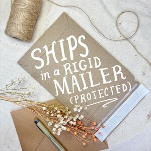 K&ouml;nnte beinhalten: Ein brauner Kartonumschlag mit dem Text "SHIPS in a RIGID MAILER (PROTECTED)" in wei&szlig; gedruckt. Der Umschlag liegt auf einer wei&szlig;en Stoffoberfl&auml;che.