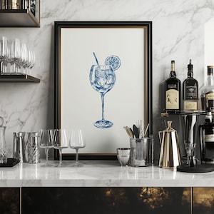 May include: A framed illustration of a blue cocktail with a lemon slice and a straw in a clear glass. The illustration is on a white background and is framed in a black frame. The frame is sitting on a countertop with other glassware and bottles.