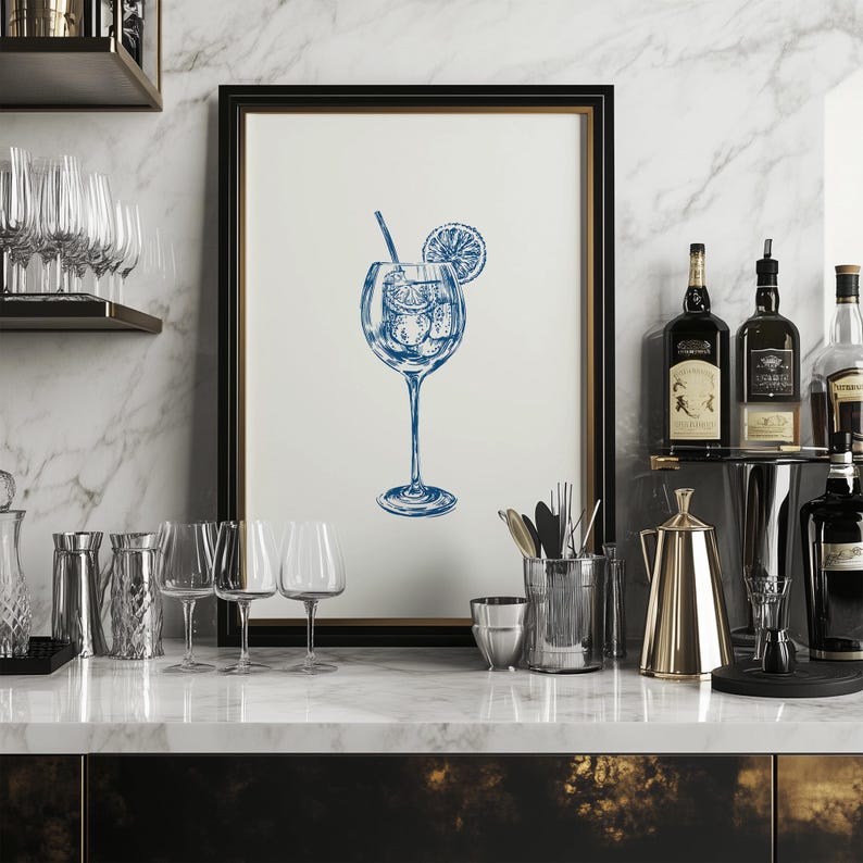 May include: A framed illustration of a blue cocktail with a lemon slice and a straw in a clear glass. The illustration is on a white background and is framed in a black frame. The frame is sitting on a countertop with other glassware and bottles.