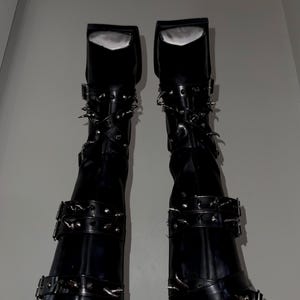May include: Pair of black knee-high boots with multiple straps, buckles, and silver-colored spikes. The boots have a square toe and a glossy finish, suggesting a leather or synthetic material. The top of the boots have a white lining.