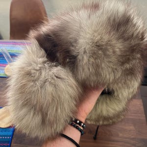 May include: A fluffy, light brown fur hat with ear flaps. The hat is held up by a person's hand. The fur is a mix of light and dark brown, creating a textured appearance. The hat has a black drawstring.