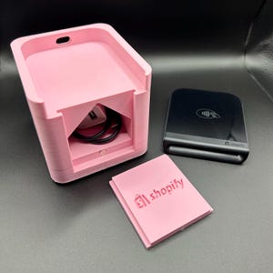May include: A pink 3D-printed Shopify card reader holder with a compartment for a charging cable. A black card reader and a pink Shopify-branded card are also present. The holder is rectangular with a slot on top.
