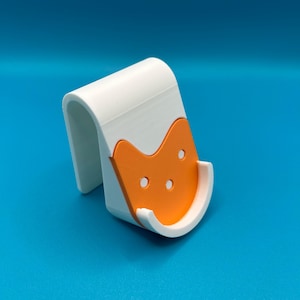 May include: White and orange phone stand with a cat face design. The stand has a curved shape and a small lip to hold a phone in place.