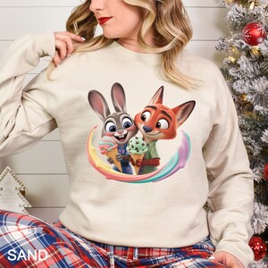 May include: Sand-colored sweatshirt featuring a colorful graphic of Zootopia characters Judy Hopps and Nick Wilde holding ice cream cones. The design includes a rainbow-colored arc. The word "SAND" is printed at the bottom.