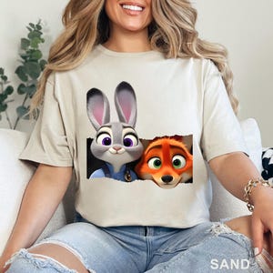 May include: Sand-colored t-shirt featuring a graphic of Judy Hopps and Nick Wilde from Zootopia. Judy, a gray rabbit, and Nick, an orange fox, are depicted peeking out from a torn-paper effect. The word "SAND" is printed at the bottom.