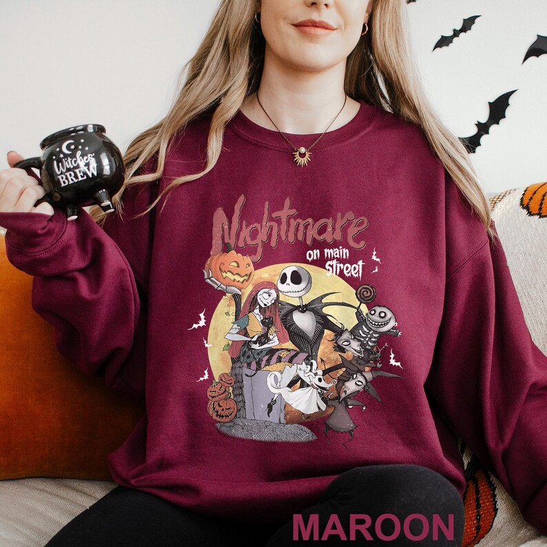 Może przedstawiać: Bordowa bluza z grafiką z filmu "Miasteczko Halloween". Projekt przedstawia Jacka Skellingtona, Sally i inne postacie na tle pełni księżyca. Tekst "Nightmare on Main Street" znajduje się nad grafiką.