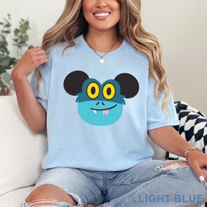 May include: Light blue t-shirt featuring a cartoon monster face with large black ears, yellow eyes, and a pink tongue. The text "LIGHT BLUE" is printed at the bottom. The shirt is a light blue color.