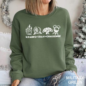 May include: Olive green sweatshirt with white graphic text and illustrations. The text reads "4 Parks 1 Day Challenge" with accompanying images of a castle, a sphere, a tree, and Mickey Mouse. The bottom right corner of the sweatshirt reads "MILITARY GREEN".