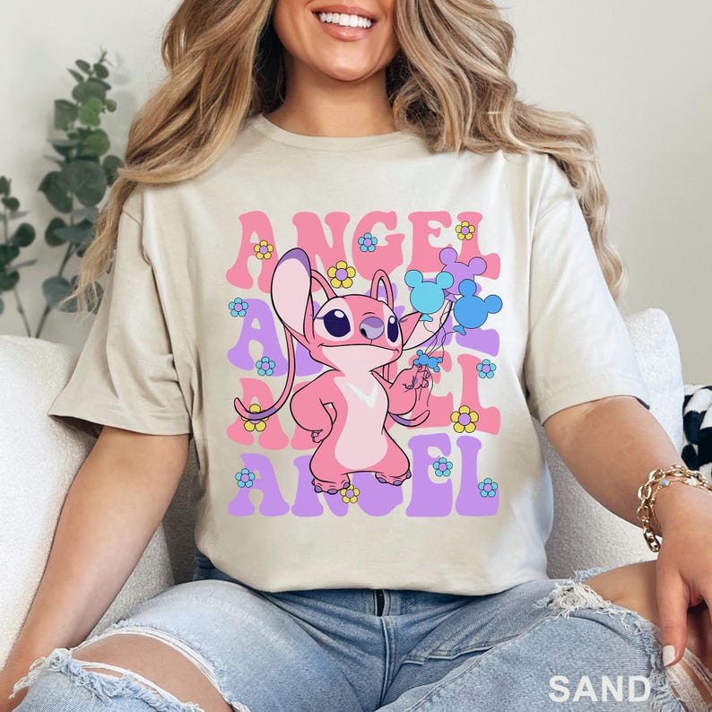 May include: Sand-colored t-shirt featuring a pink cartoon character holding blue balloons. The word "ANGEL" is printed in pink and purple, with flower accents. The shirt is a casual wear item.