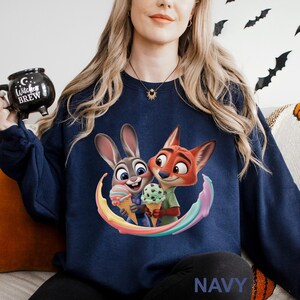 May include: Navy blue sweatshirt featuring a colorful graphic of Judy Hopps and Nick Wilde from Zootopia holding ice cream cones. The word "NAVY" is printed at the bottom. The sweatshirt is a comfortable, casual style.