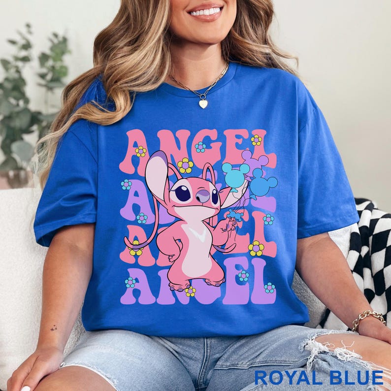 May include: Royal blue t-shirt featuring a pink cartoon character holding Mickey Mouse-shaped balloons. The word "ANGEL" is printed vertically in pink and lavender, with flower and Mickey Mouse accents.