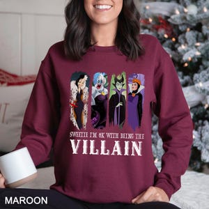May include: Maroon sweatshirt featuring four cartoon villain characters. The text "Sweetie I'm OK with being the Villain" is printed below the characters in white. The word "MAROON" is printed at the bottom.
