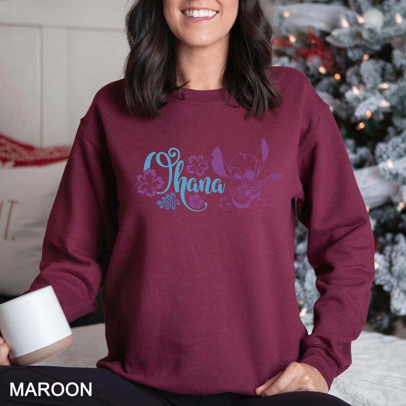 May include: Maroon sweatshirt featuring the word "Ohana" in teal script with floral accents and a purple cartoon character playing a ukulele. The sweatshirt is a solid maroon color.
