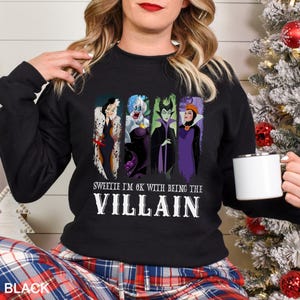 May include: Black sweatshirt featuring four cartoon villain characters. The text "Sweetie I'm OK with being the Villain" is printed below the characters in white. The word "BLACK" is printed at the bottom left.