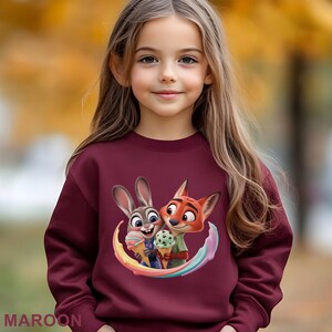 May include: A maroon sweatshirt featuring a colorful graphic of the Zootopia characters Judy Hopps and Nick Wilde holding ice cream cones. The word "MAROON" is printed at the bottom. The sweatshirt is worn by a young girl.