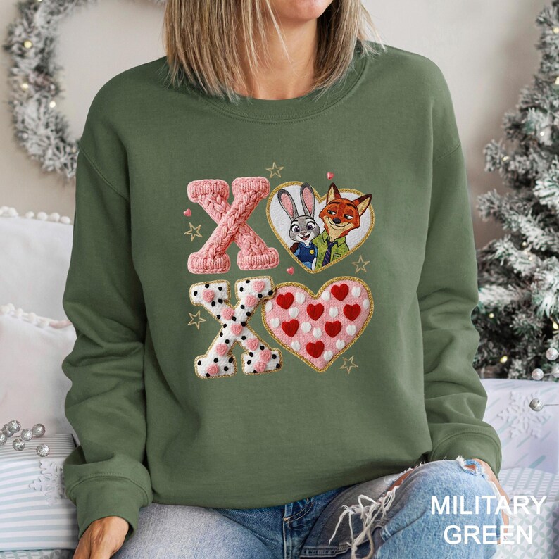 May include: Military green sweatshirt with embroidered design. Features the letters "XO" with a heart-shaped design and cartoon characters. The top letter "X" is pink knit, the "O" is a heart with cartoon characters, and the bottom "X" is white with pink polka dots.