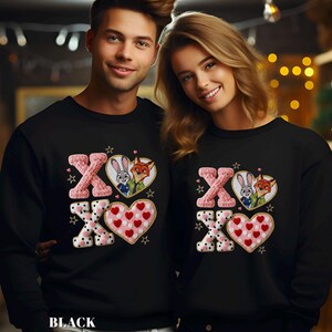May include: Black sweatshirts with a Valentine's Day design. The design includes the letters "XO", hearts, and images of cartoon characters. The sweatshirts are worn by two people.