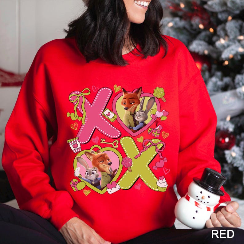 Peut inclure: Un sweat-shirt rouge avec un motif repr&eacute;sentant les personnages de Zootopie, Nick Wilde et Judy Hopps. Le motif comprend des formes X et O roses et vertes, des c&oelig;urs et d'autres graphiques sur le th&egrave;me de la Saint-Valentin. Le mot "RED" est imprim&eacute; en blanc en bas &agrave; droite.