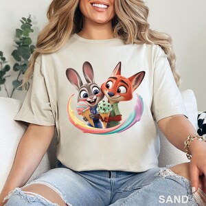 May include: Sand-colored t-shirt featuring a colorful graphic of Judy Hopps and Nick Wilde from Zootopia, each holding ice cream cones. The design includes a rainbow-colored swirl. The word "SAND" is printed at the bottom.
