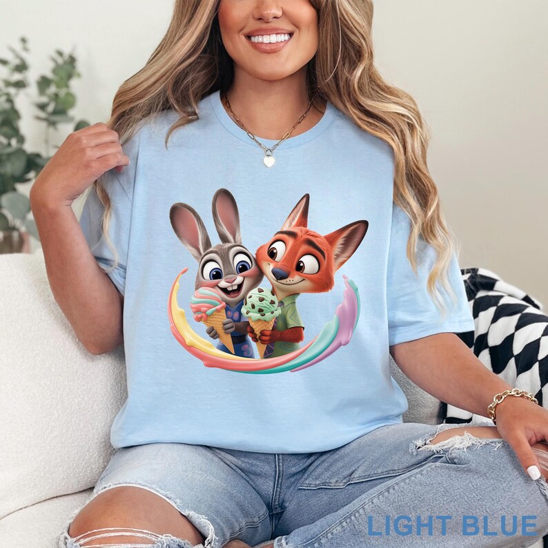 May include: Light blue t-shirt featuring a colorful graphic of two cartoon characters, a rabbit and a fox, holding ice cream cones. The shirt has the words "LIGHT BLUE" printed at the bottom.