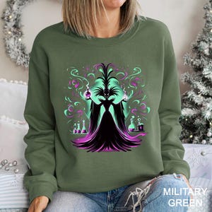 Op de afbeelding: Militairgroen sweatshirt met een afbeelding van een donkere figuur in een zwarte jurk en een turquoise gezicht. De figuur is omgeven door wervelende turquoise en paarse rook, met laboratoriumflessen aan weerszijden. De tekst "MILITARY GREEN" staat rechtsonder.