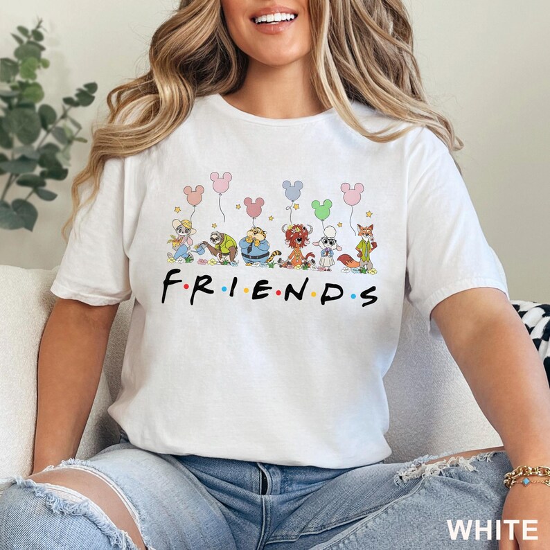 Puede incluir: Camiseta blanca con la palabra "FRIENDS" en letras negras. Encima de la palabra, personajes de dibujos animados sostienen globos con forma de Mickey Mouse. La camiseta est&aacute; hecha de un material suave y es una prenda informal.