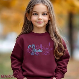 May include: Maroon sweatshirt featuring the word "Ohana" in a stylized font with floral accents and a purple cartoon character playing a guitar. The word "MAROON" is printed at the bottom of the image.