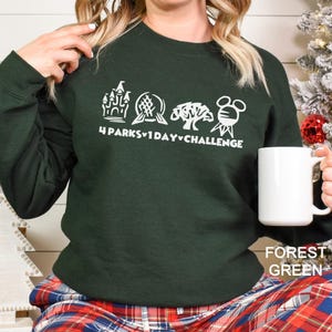 May include: Forest green sweatshirt with a white graphic of four theme park icons and the text "4 Parks 1 Day Challenge." A white mug is held in the hand. The sweatshirt is paired with plaid pajama pants.