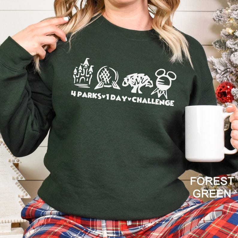 May include: Forest green sweatshirt with a white graphic of four theme park icons and the text "4 Parks 1 Day Challenge." A white mug is held in the hand. The sweatshirt is paired with plaid pajama pants.