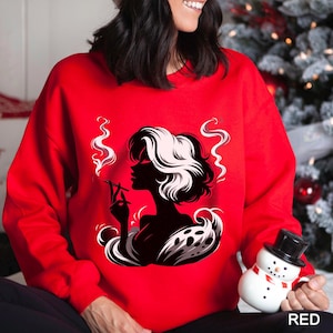 Op de afbeelding: Een rode sweatshirt met een zwart-witte afbeelding van een rokende vrouw. Het silhouet van de vrouw is in profiel, met tweekleurig haar. Het woord "RED" staat rechtsonder op de sweatshirt.