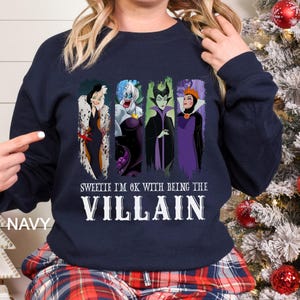 May include: Navy blue sweatshirt featuring four cartoon villains: Cruella de Vil, Ursula, Maleficent, and the Evil Queen. The text "Sweetie I'm OK with being the Villain" is printed below the characters in white. The word "NAVY" is printed on the left side.
