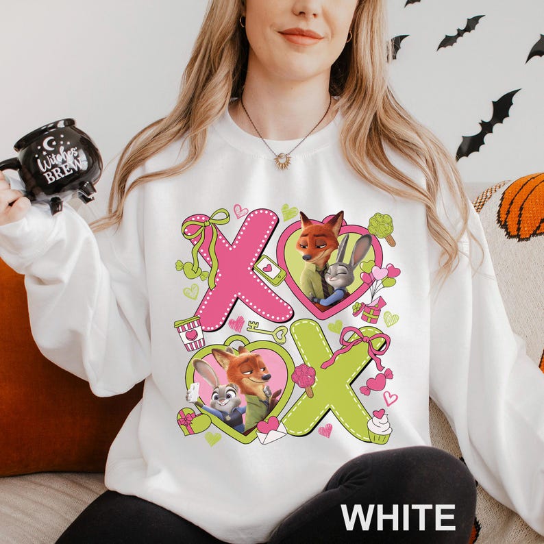 Peut inclure: Sweat-shirt blanc avec un motif color&eacute; des personnages de Zootopie, Nick Wilde et Judy Hopps. Le design comprend des X roses et verts, des c&oelig;urs et d'autres illustrations. Le mot "WHITE" est imprim&eacute; en bas.