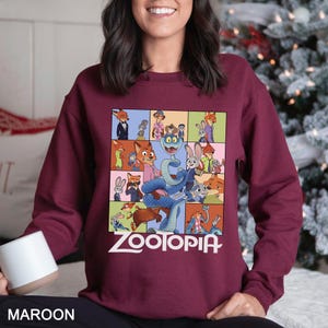 May include: Maroon sweatshirt featuring a colorful graphic of characters from the animated film "Zootopia." The design includes a grid of images with the film's title in white. The sweatshirt is a solid maroon color.