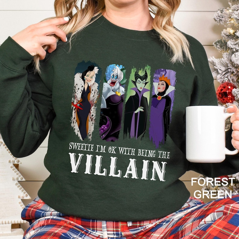 May include: Forest green sweatshirt featuring four cartoon villain characters. The text "Sweetie I'm OK with being the Villain" is printed below the characters in white. The sweatshirt is a long-sleeved crew neck.