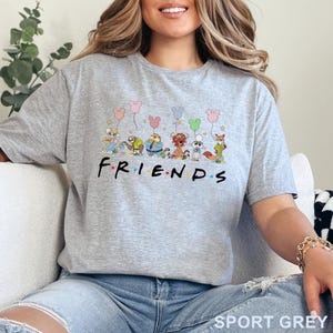 Puede incluir: Camiseta gris deportiva con la palabra "FRIENDS" en negro y puntos de colores. Encima de la palabra, personajes de dibujos animados sostienen globos con forma de Mickey Mouse. La camiseta es de un material suave.