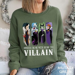 May include: Military green sweatshirt featuring four cartoon villain characters. The text "Sweetie I'm OK with being the Villain" is printed below the characters in white. The sweatshirt is a crew neck style.