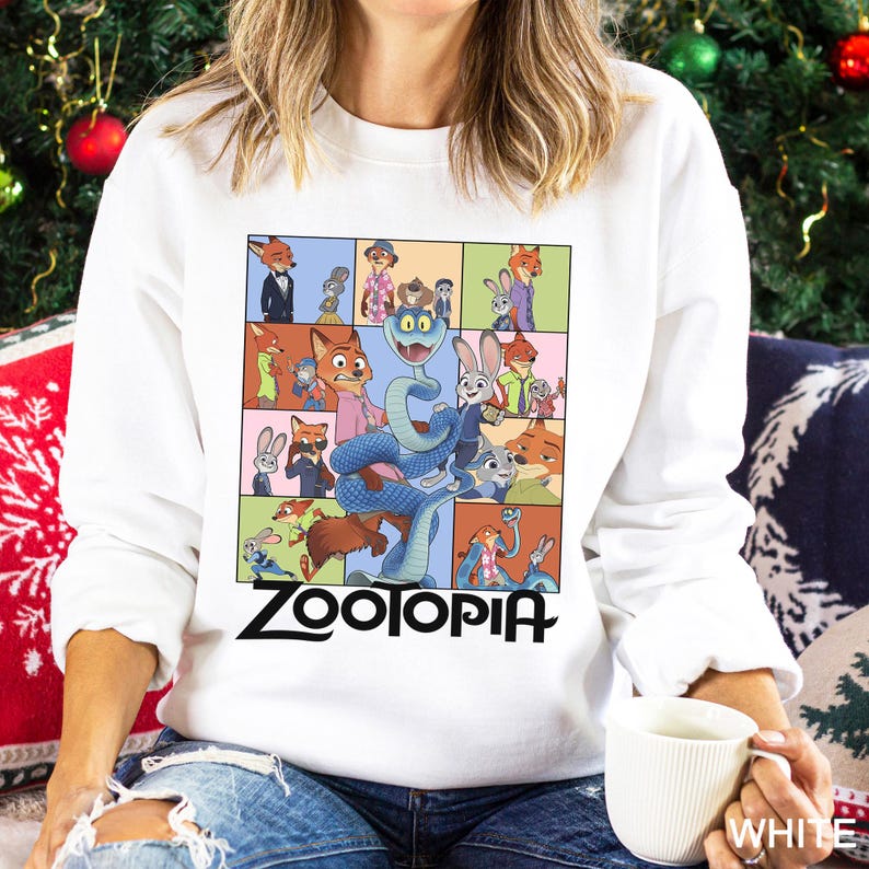 May include: White sweatshirt featuring a colorful grid of Zootopia characters, including a blue snake and various animal figures. The word "ZOOTOPIA" is printed in black block letters at the bottom. The sweatshirt is worn by a person.