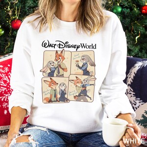 May include: White sweatshirt with a Walt Disney World graphic. The design features cartoon images of Nick Wilde and Judy Hopps, with the phrases "Sweet Surrender", "Adventure Awaits", and "Dancing Through Life". The sweatshirt is made of a soft material.