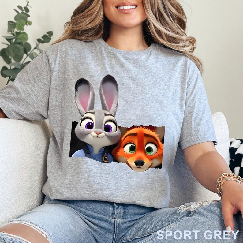 May include: Sport grey t-shirt featuring a graphic of Judy Hopps and Nick Wilde from Zootopia. The characters are depicted in a torn pocket design. The shirt is made of a soft material.