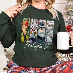 Puede incluir: Sudadera verde bosque con personajes de Zootopia 2. El dise&ntilde;o incluye a Nick Wilde, Judy Hopps, el jefe Bogo y Flash. El texto "Zootopia 2" est&aacute; escrito en blanco. La sudadera es de color liso.