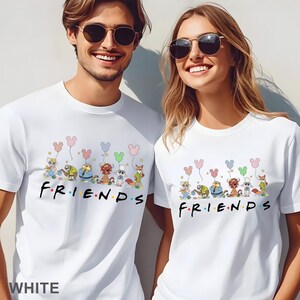 Puede incluir: Camisetas blancas con un dise&ntilde;o de dibujos animados de personajes que sostienen globos con forma de Mickey Mouse sobre la palabra "FRIENDS" en letras coloridas. Las camisetas son usadas por dos personas.