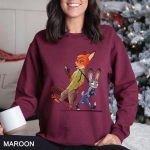 May include: Maroon sweatshirt featuring a cartoon illustration of a fox and a rabbit. The fox is holding a phone and the rabbit is wearing a police uniform. The word "MAROON" is printed at the bottom of the sweatshirt.