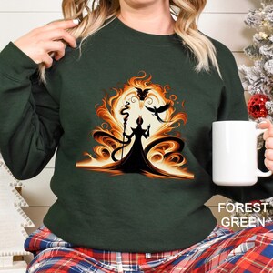 Op de afbeelding: Boszwarte groene sweatshirt met een afbeelding van een silhouet met een staf, tegen een vurige oranje en gele achtergrond. Een zwarte vogel vliegt in de buurt. De tekst "FOREST GREEN" staat rechtsonder.